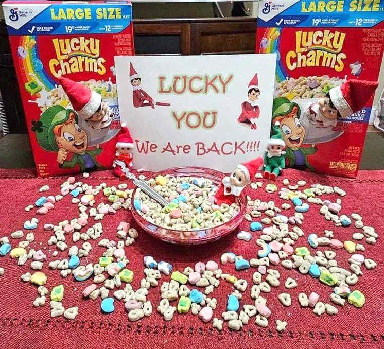 A bowl of Lucky Charms cereal is surrounded by scattered marshmallows, boxes of Lucky Charms, Elf on the Shelf dolls, and a sign that says, โLUCKY YOU, We Are BACK!!!!โ on a red tablecloth.