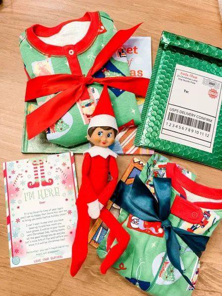 A festive display includes an Elf on the Shelf doll, two green Christmas pajamas tied with red and blue ribbons, a holiday-themed card, a book, and a green USPS padded envelope on a wooden surface.