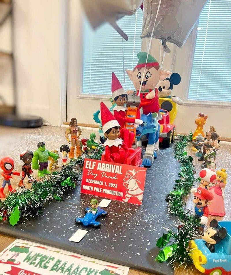 A festive display features elves in a sleigh pulled by toy vehicles, surrounded by various action figures and dolls. A sign reads โElf Arrivalโ with the date December 1, 2023, and holiday decorations line the scene.