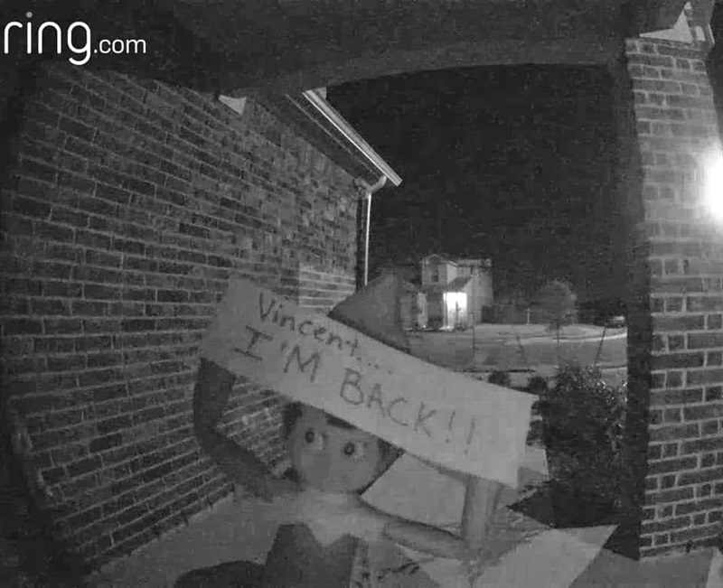 A night-time doorbell camera image shows an elf doll holding a sign that reads "Vincent... I'M BACK!!" in front of a brick house.