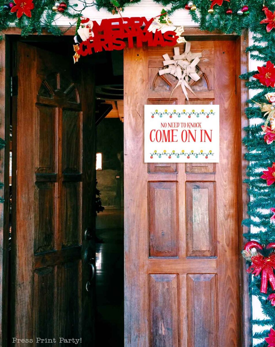 A wooden door decorated with a "Merry Christmas" sign, garland, and red flowers stands open. A sign on the door reads, "No need to knock, come on in" in festive red and green letters.