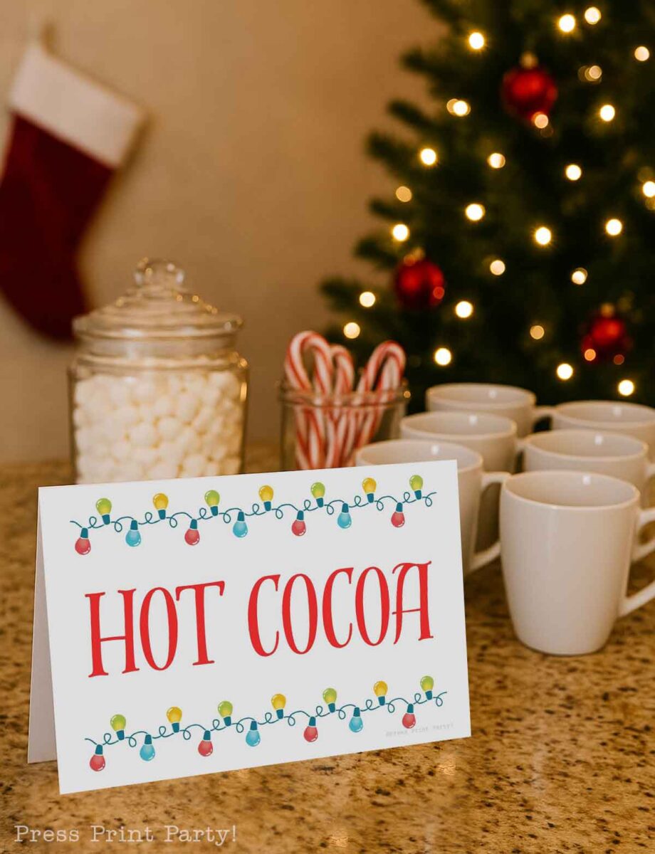A table with a "HOT COCOA" sign, mugs, a jar of marshmallows, a jar of candy canes, and a decorated Christmas tree with lights and red ornaments in the background.