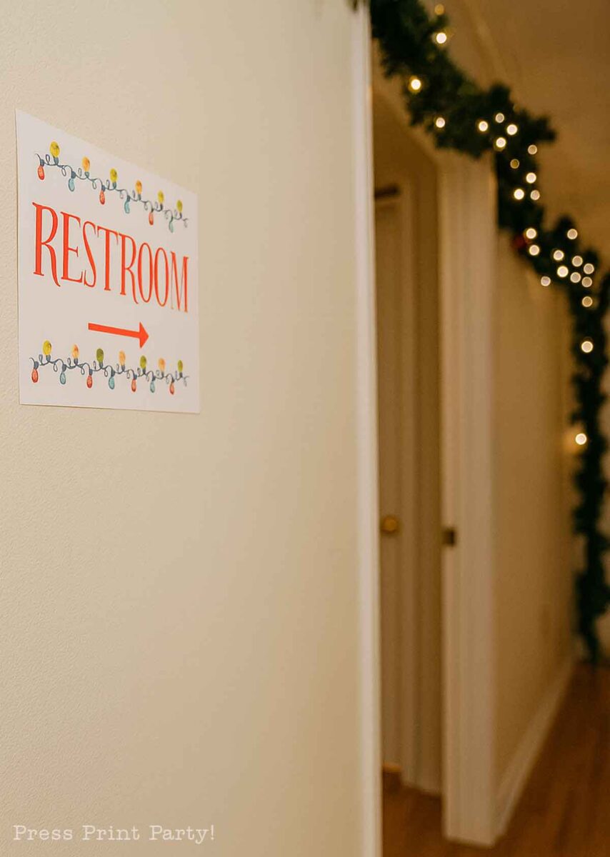 A wall sign with the word "Restroom" and a right-pointing red arrow, decorated with colorful string lights, directs toward a hallway adorned with holiday garlands and string lights.