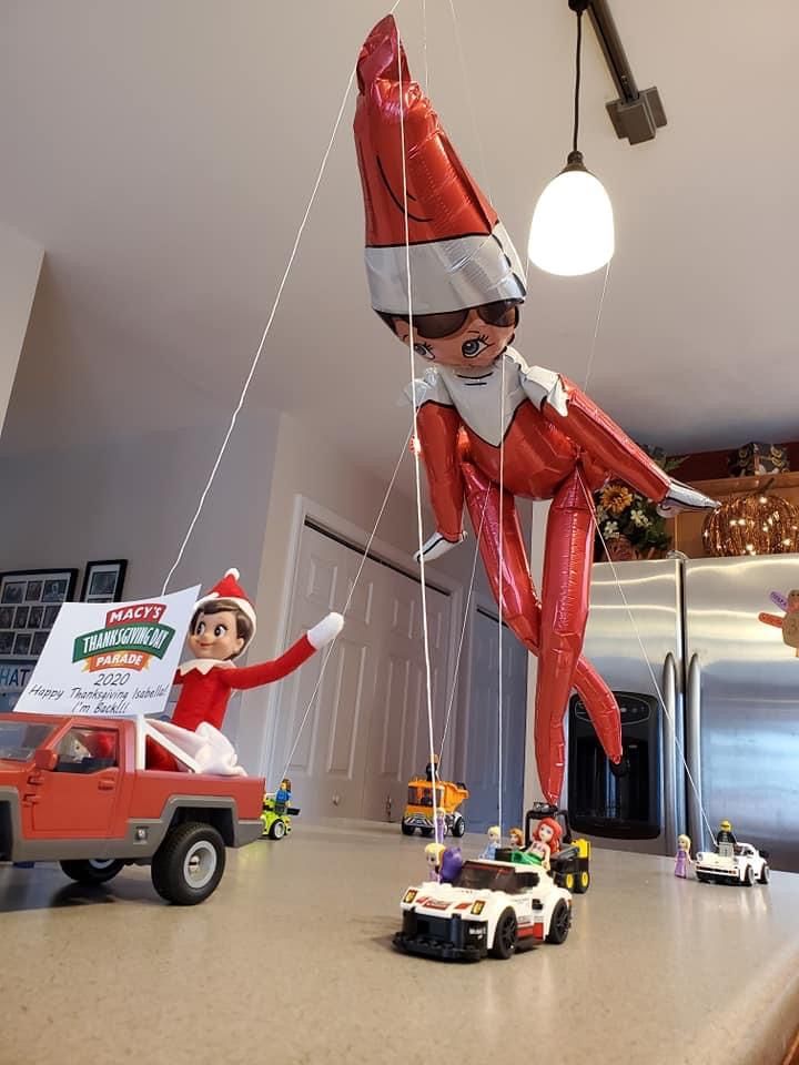 A balloon elf on the shelf is suspended above a kitchen counter by strings, while a toy elf in a red truck leads small toy cars, recreating a parade scene. The background features a fridge and kitchen decor.