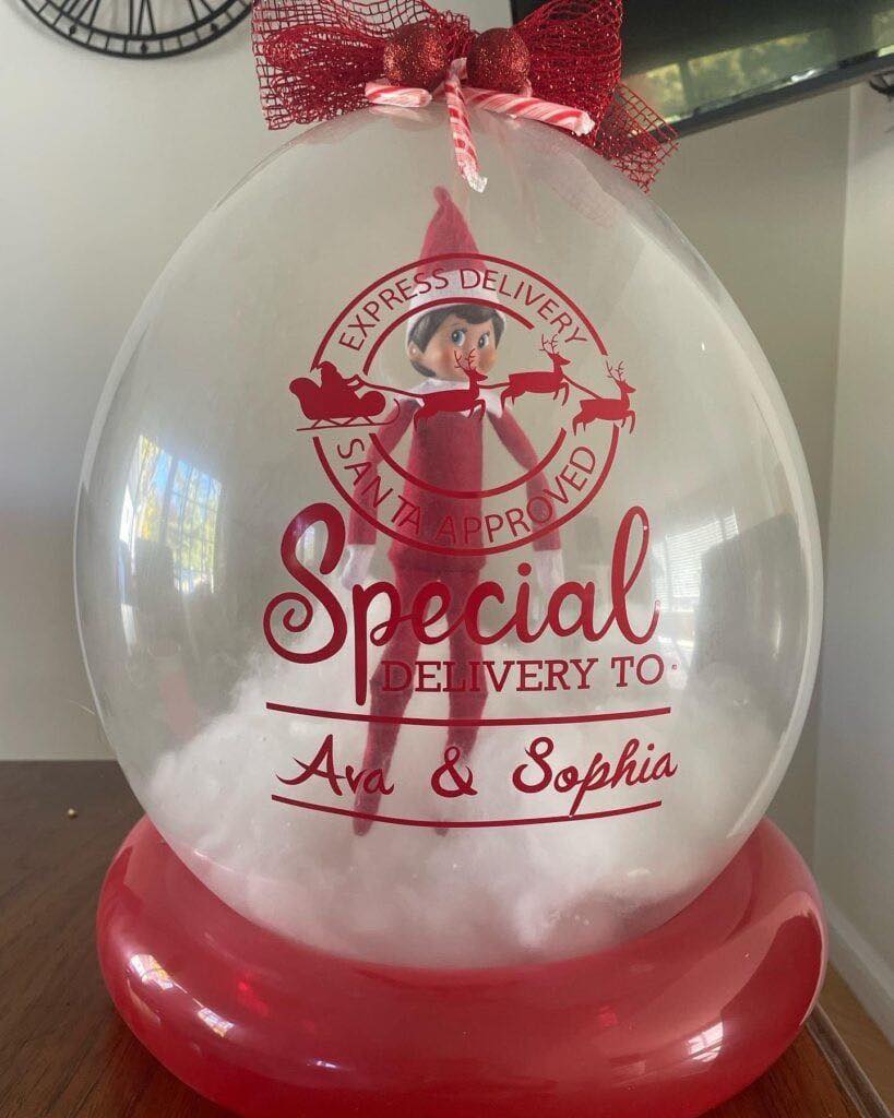 A transparent balloon with "Special Delivery to Ava & Sophia" and Santa-themed graphics contains an Elf on the Shelf doll inside, resting on artificial snow, topped with a red bow and set on a red balloon base.
