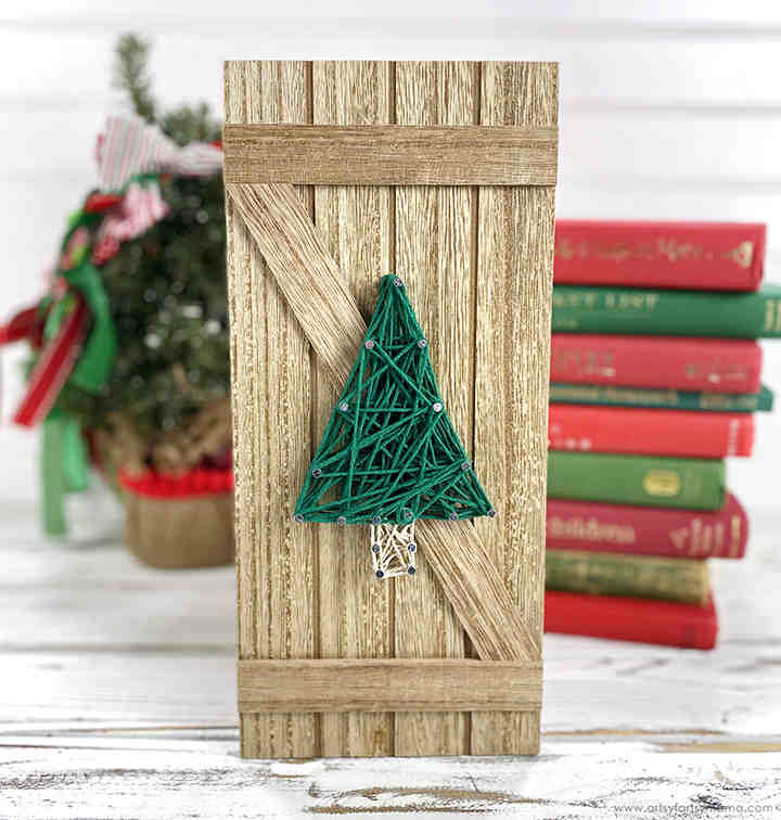 A rustic wooden plaque decorated with green string art in the shape of a Christmas tree stands on a white surface. In the background are stacked books and a small decorated Christmas tree.