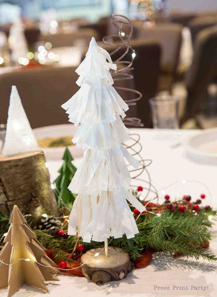 A festive table centerpiece featuring a white, tiered, paper Christmas tree with a spiral wire topper, surrounded by greenery, red berries, and other small decorative trees.