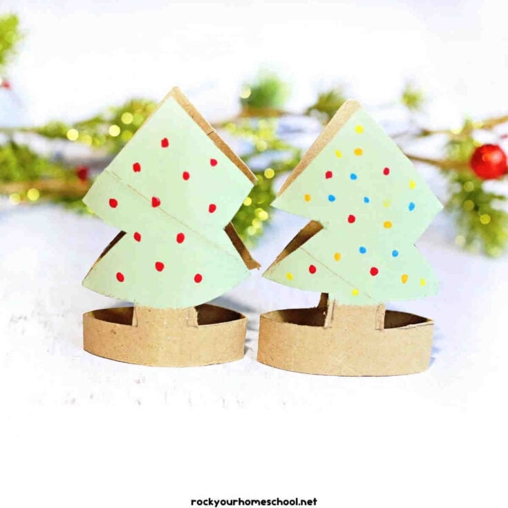 Two small Christmas tree crafts made from green paper and cardboard, decorated with colorful dots, stand side by side with blurred greenery and red berries in the background.