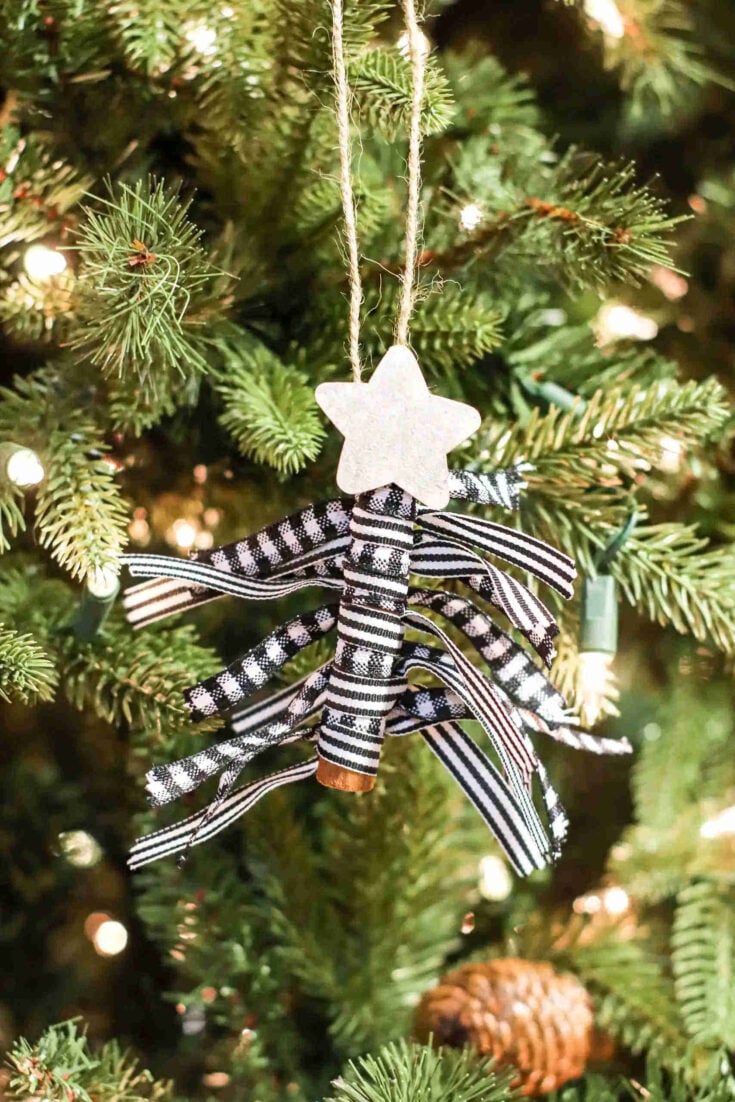 A handmade Christmas tree ornament with a white star on top, black and white ribbon strips as branches, and a cinnamon stick as the trunk, hangs on a green Christmas tree with lights.