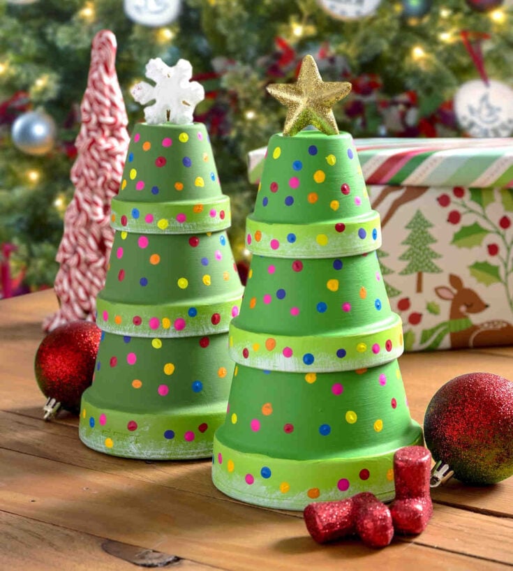 Two small Christmas trees made from stacked, painted green flower pots decorated with colorful dots, topped with a star and snowflake, sit on a wooden table with ornaments and a wrapped gift in front of a Christmas tree.