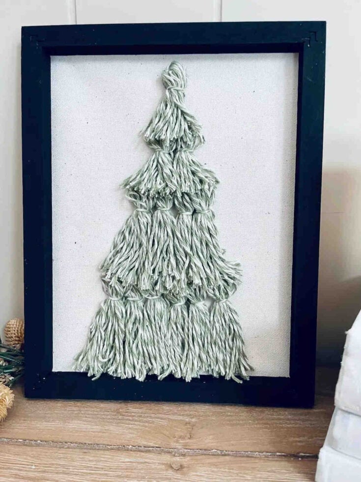 Framed artwork featuring a textured Christmas tree made from light green yarn on a white canvas, set in a black frame and displayed on a wooden surface.