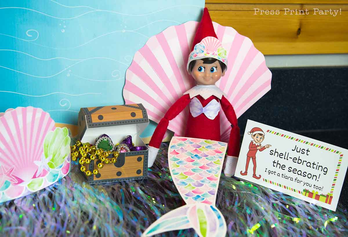 A festive Elf on the Shelf dressed as a mermaid sits between paper seashells and printable Elf on the Shelf props, next to a treasure chest filled with jewels and a sign that says, "Just shell-ebrating the season! I got a tiara for you too!.