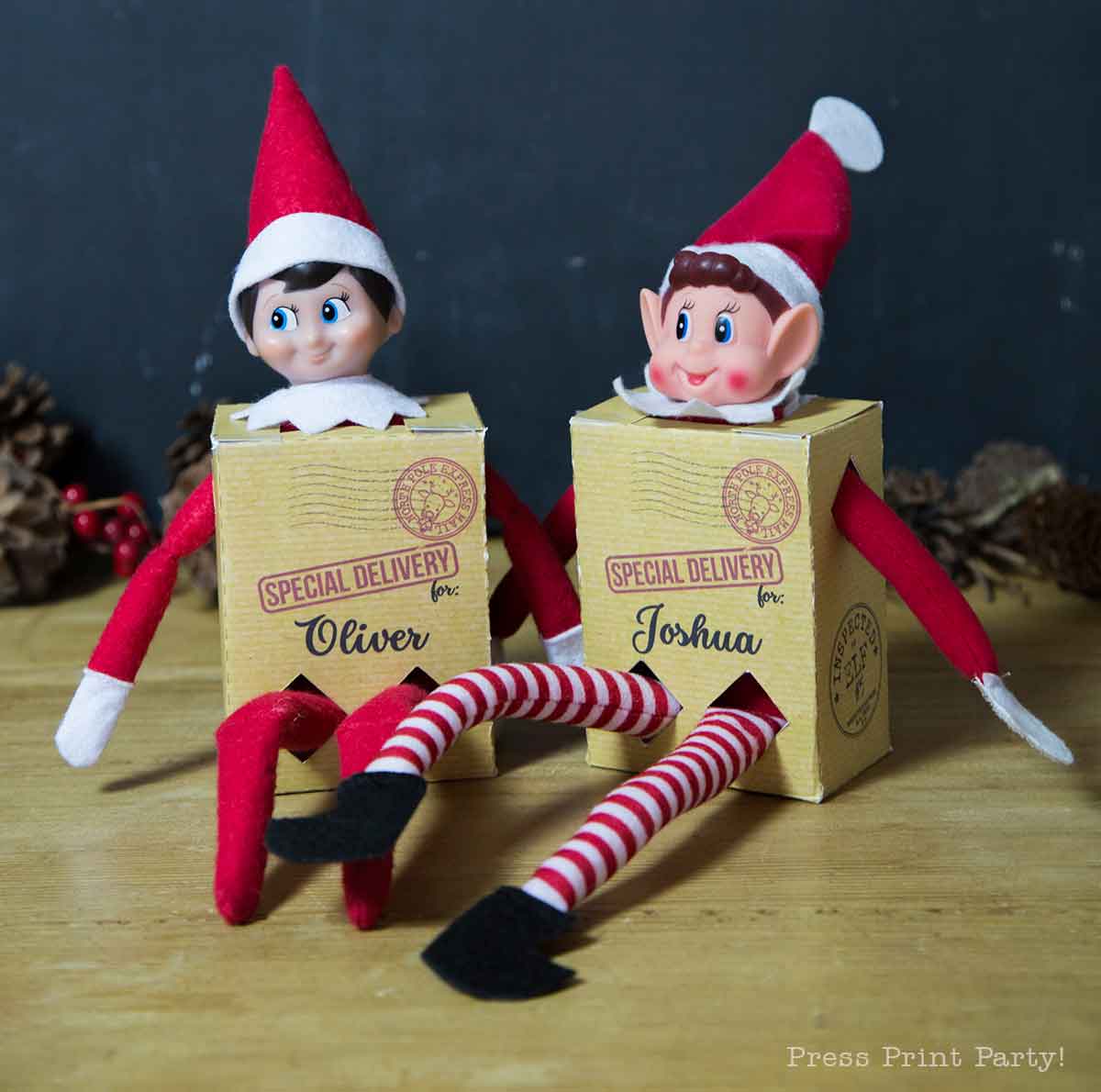 Two Elf on the Shelf dolls sit with their arms and legs outstretched, each inside a small box labeled "Special Delivery for Oliver" and "Special Delivery for Joshua," featuring printable Elf on the Shelf props, on a wooden surface with pinecones in the background.