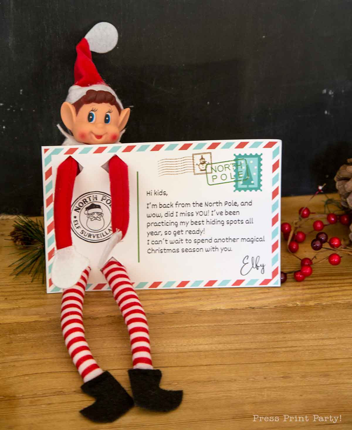 A toy elf with a red hat and striped legs sits on a table, holding a note from the North Pole. Festive decorations and a dark wall set the scene, while printable elf on the shelf props add extra holiday fun.