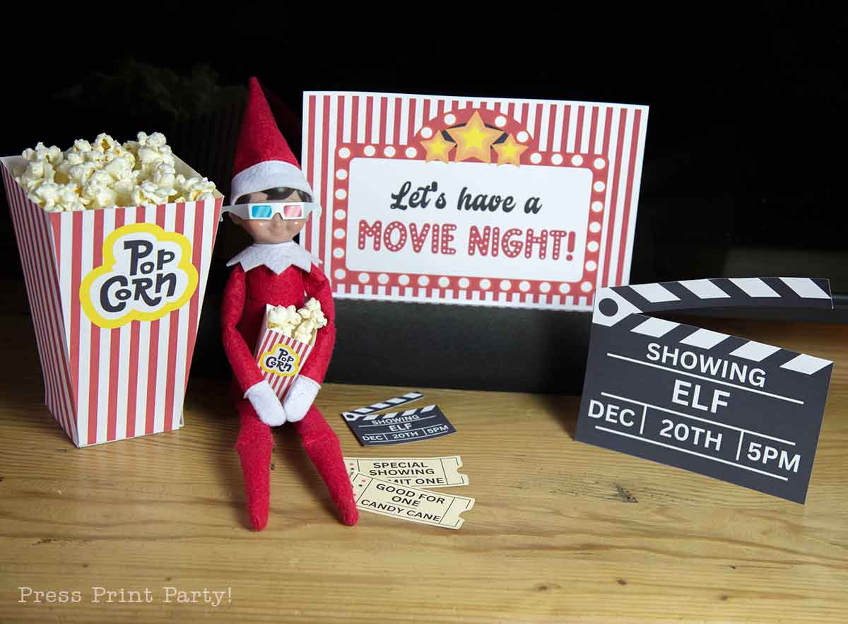 A plush elf doll wearing 3D glasses sits with a popcorn box, surrounded by movie night decorations, printable Elf on the Shelf props, a clapperboard, and movie snacks—a perfect setup for "Let's have a Movie Night!" fun.