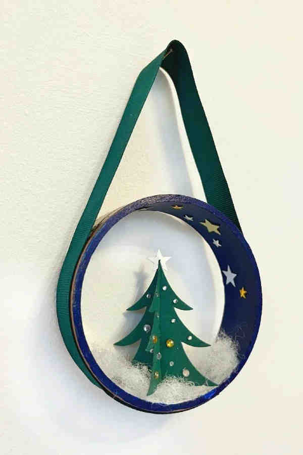A handmade ornament featuring a small green paper Christmas tree with decorations, set inside a blue embroidery hoop with star stickers, white fluff as snow, and a green ribbon for hanging.