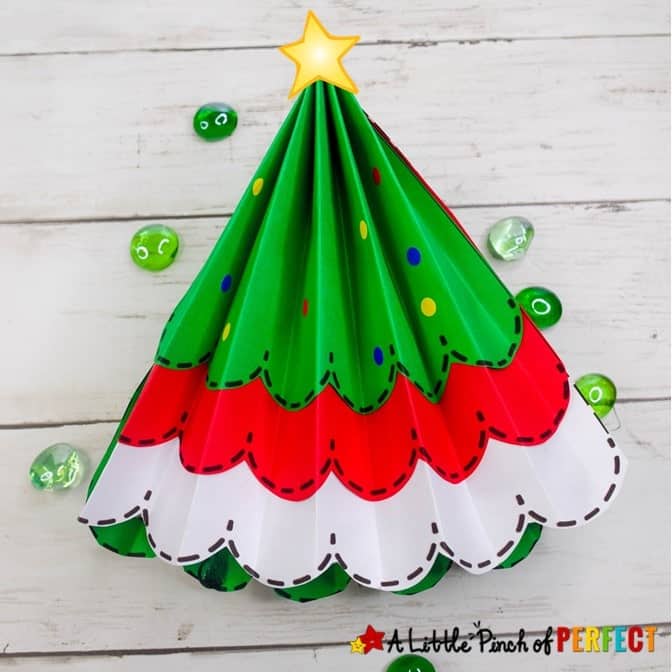 Folded paper Christmas tree craft with green, red, and white scalloped layers, decorated with dots and a yellow star on top, placed on a white wooden surface with green glass pebbles around it.