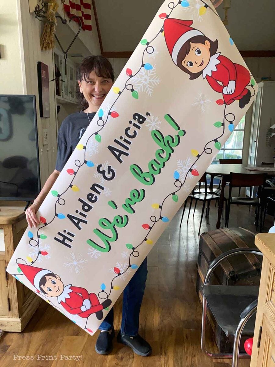 A smiling woman holds a large festive banner that reads "Hi Aiden & Alicia! We’re back!" decorated with cartoon elves, printable elf on the shelf props, snowflakes, and colorful holiday lights.