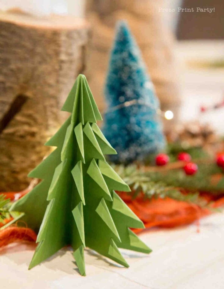 A close-up of a green paper origami Christmas tree, with a blue bottle brush tree and festive decorations like red berries and greenery blurred in the background.
