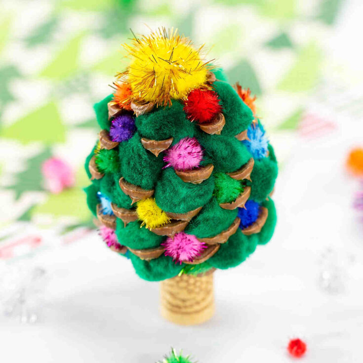 A handmade Christmas tree craft made from green pom-poms, walnut shell halves, and colorful mini pom-poms, topped with a gold tinsel pom-pom, standing on a cork base. The background is blurred and festive.