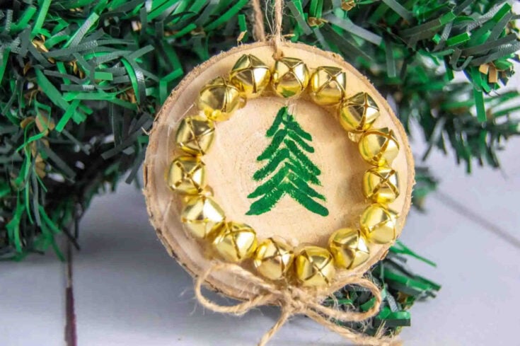 A wooden ornament painted with a green Christmas tree, decorated with a circle of small gold bells and tied with twine, hangs in front of artificial pine branches.