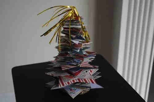 A small Christmas tree decoration made from stacked colorful, patterned paper squares, topped with shiny gold tinsel, sits on a dark surface near a window.