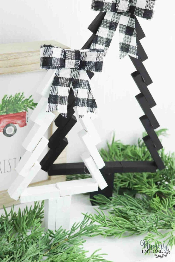 Two Christmas tree decorations made from painted clothespins, one white and one black, each topped with a black-and-white checkered bow, sit among green faux pine branches.