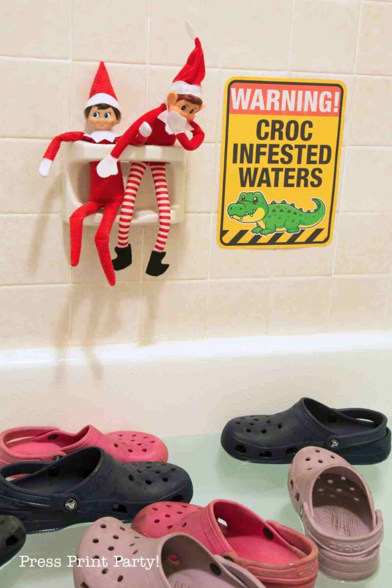 Two Elf on the Shelf dolls perch on a bathtub soap holder above a "Warning! Croc Infested Waters" sign, with printable elf on the shelf props and pairs of Crocs shoes floating in the tub water below.