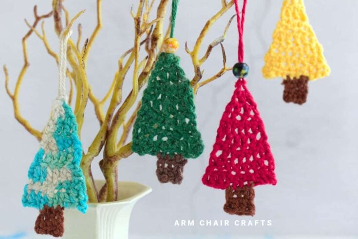 Four colorful crocheted Christmas tree ornaments hang from the branches of a bare decorative tree in a white vase. The trees are in blue, green, red, and yellow with brown bases.