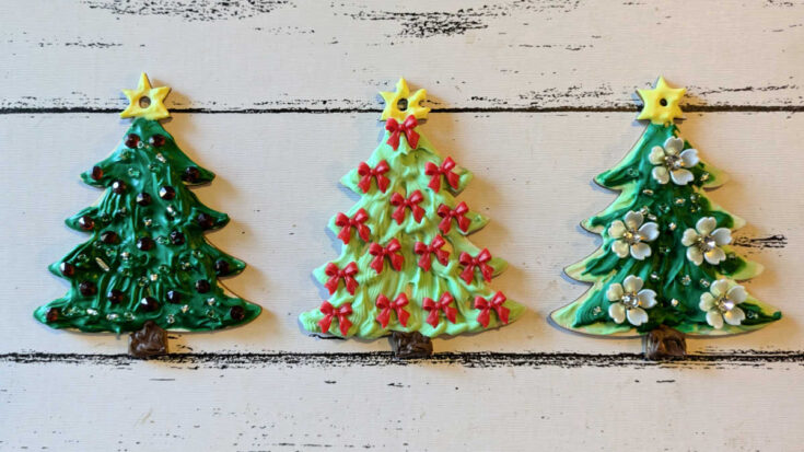 Three decorated Christmas tree cookies are arranged in a row on a white wooden surface, each with a yellow star on top and unique decorations: red ornaments, red bows, and white flowers.