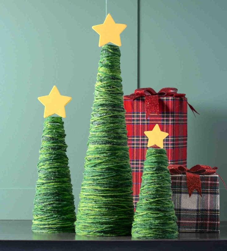 Three cone-shaped Christmas trees made of green yarn, each topped with a yellow star, stand in front of two red and black plaid gift boxes with shiny red bows, against a green background.