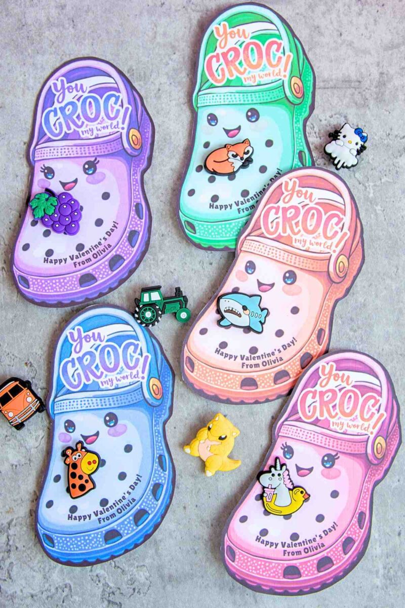 Five colorful Valentine's Day cards shaped like Crocs shoes are decorated with cute animal illustrations and the text "You Croc my world! Happy Valentine's Day! From Olivia." Small toy pins surround the cards.