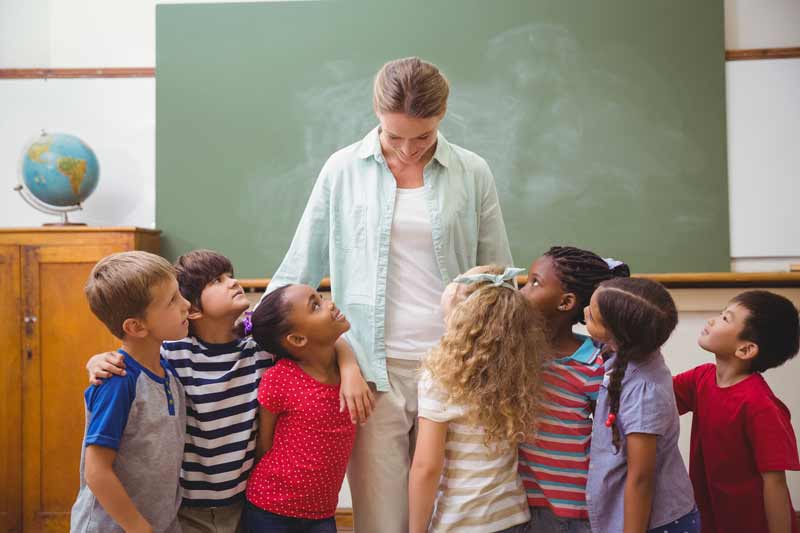 A teacher stands in front of a chalkboard, smiling down at a diverse group of young children who are gathered around her, eager to start some fun ice breaker games for kids. A globe sits on a cabinet in the background.