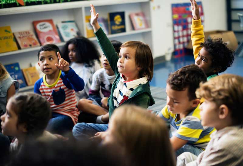 A group of young children sit on the floor in a classroom, with bookshelves behind them. Two children in the center have their hands raised, eager to participate in fun ice breaker games for kids. The atmosphere is lively and engaged.