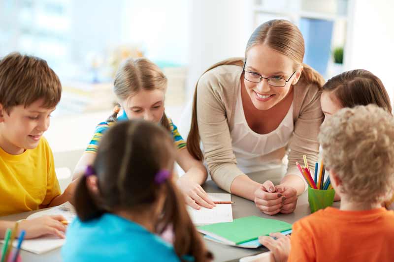 A teacher with glasses smiles and interacts with a group of young children sitting around a table, working with papers and colored pencils in a bright classroom, as they enjoy fun ice breaker games for kids.