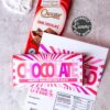 A Valentine’s Day chocolate bar with a pink and white wrapper sits on a card with custom text, next to the Valentines Day Socks Wrap Printable, another Choceur dark chocolate bar, and a crocheted heart.