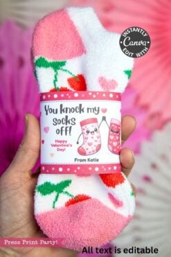 A hand holds a pair of fuzzy socks decorated with strawberries and hearts, wrapped in the "Valentines Day Socks Wrap Printable" that says, "You knock my socks off! Happy Valentine's Day! From Katie," with two cartoon smiling socks.