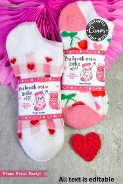 Two pairs of fuzzy Valentine’s socks—pink plaid and white with strawberries—are wrapped with the Valentines Day Socks Wrap Printable and displayed next to Kinder Bueno Valentines on a gray surface, with festive red and pink decor in the background.