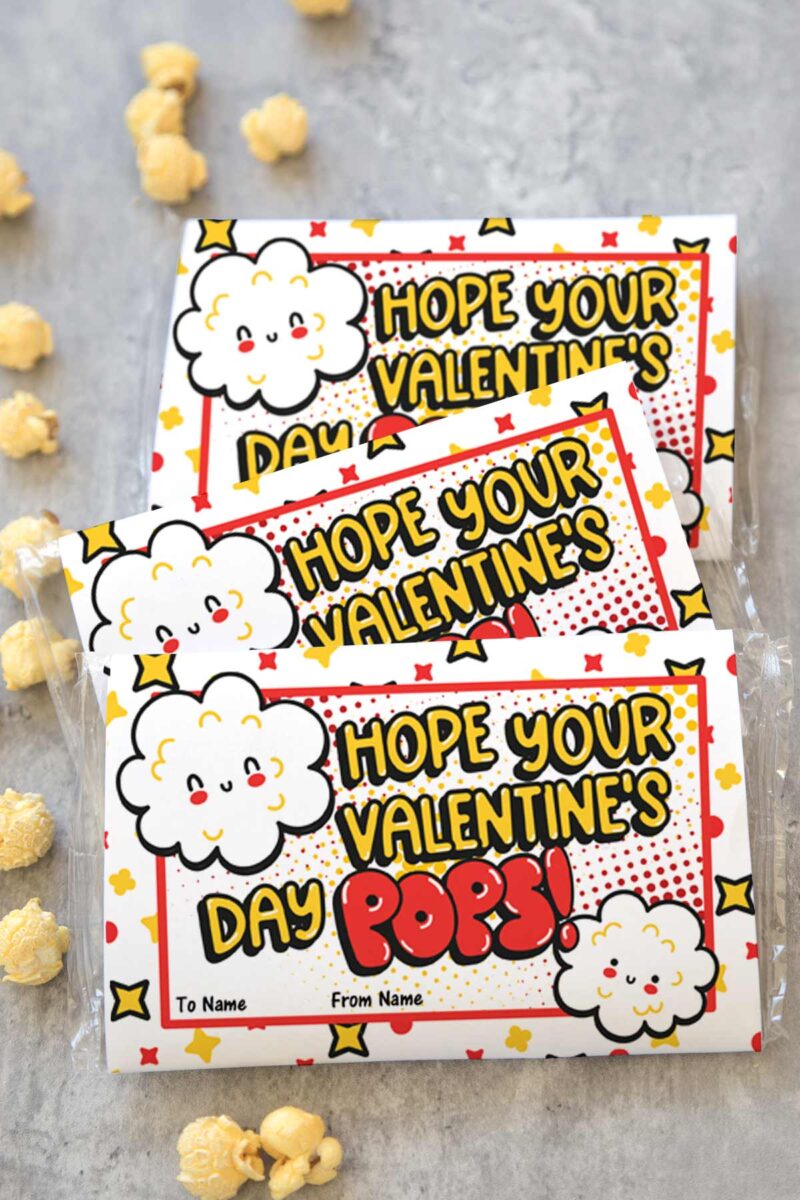 Three popcorn snack bags with colorful labels featuring smiling popcorn illustrations and the message "Hope your Valentine's Day POPS!" There are spots to write "To" and "From" names. Pieces of popcorn are scattered around.