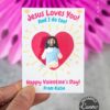 A hand holds the "Jesus Loves You Valentine Card," showing a clay Jesus inside a red heart. The card says, “Jesus Loves You! And I do too! Happy Valentine’s Day! From Katie.” Blurred pink decor and panda party favors are in the background.