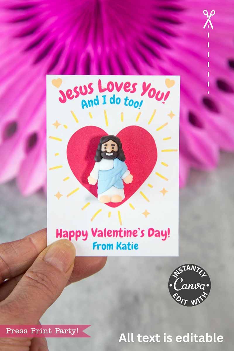 A hand holds the "Jesus Loves You Valentine Card," showing a clay Jesus inside a red heart. The card says, “Jesus Loves You! And I do too! Happy Valentine’s Day! From Katie.” Blurred pink decor and panda party favors are in the background.