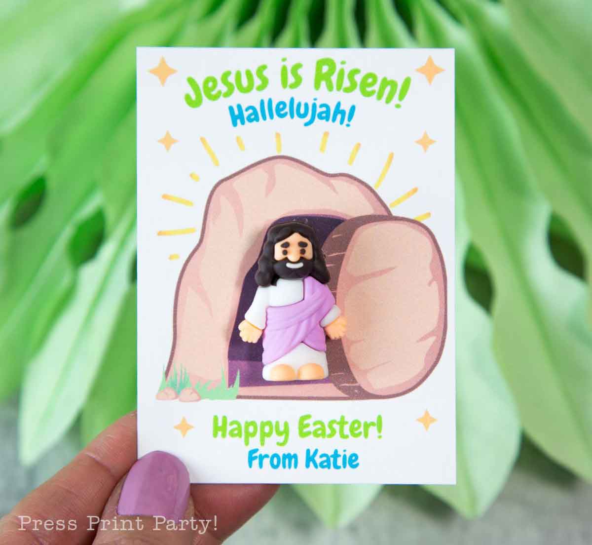 A hand holds a card with an illustration of the risen Jesus standing in front of an empty tomb. The card reads, "Jesus is Risen! Hallelujah! Happy Easter! from Katie" with leaves in the background.