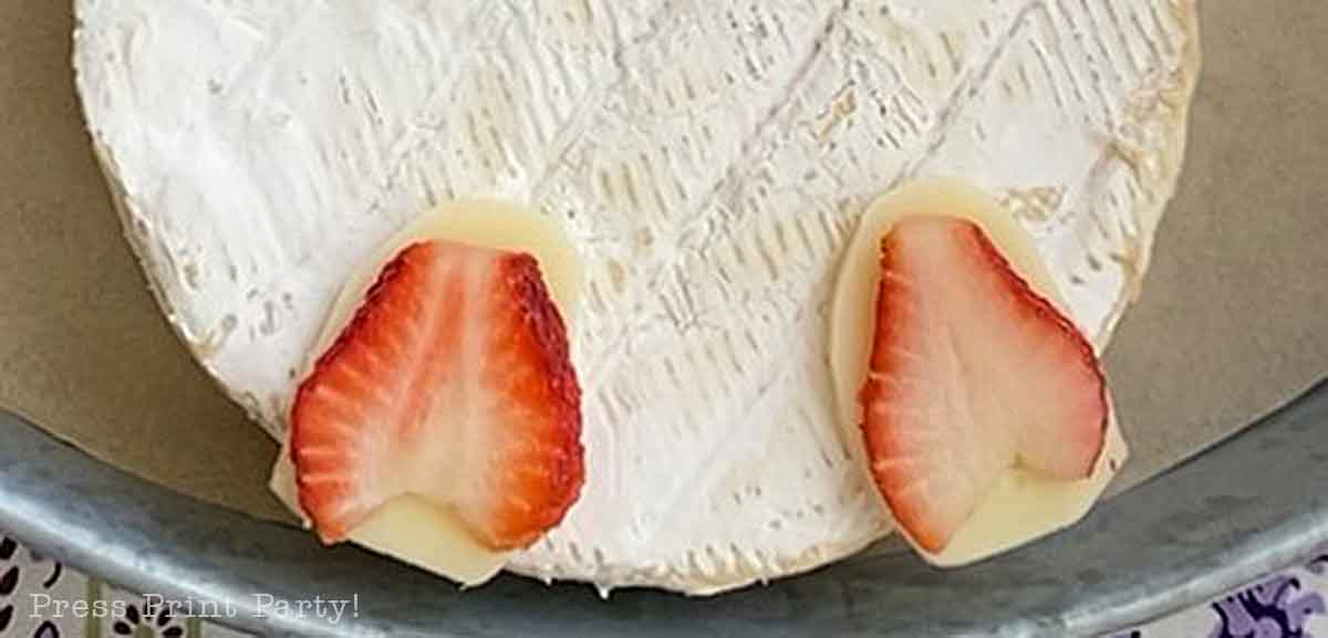 A round wheel of brie cheese with two thin slices of strawberry on top, resembling bunny earsβperfect for adding a playful touch to your Easter Bunny Charcuterie Board.