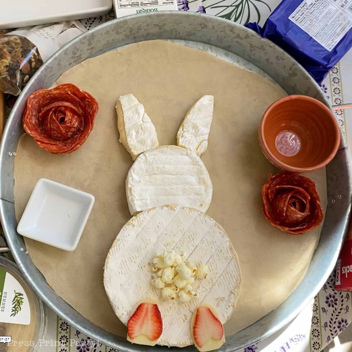 A whimsical Easter Bunny Charcuterie Board features a cheese platter shaped as a bunny, with cheese wheels for the body and head, cheese ears, popcorn tail, strawberry feet, meat roses, and small bowls arranged on a round tray.