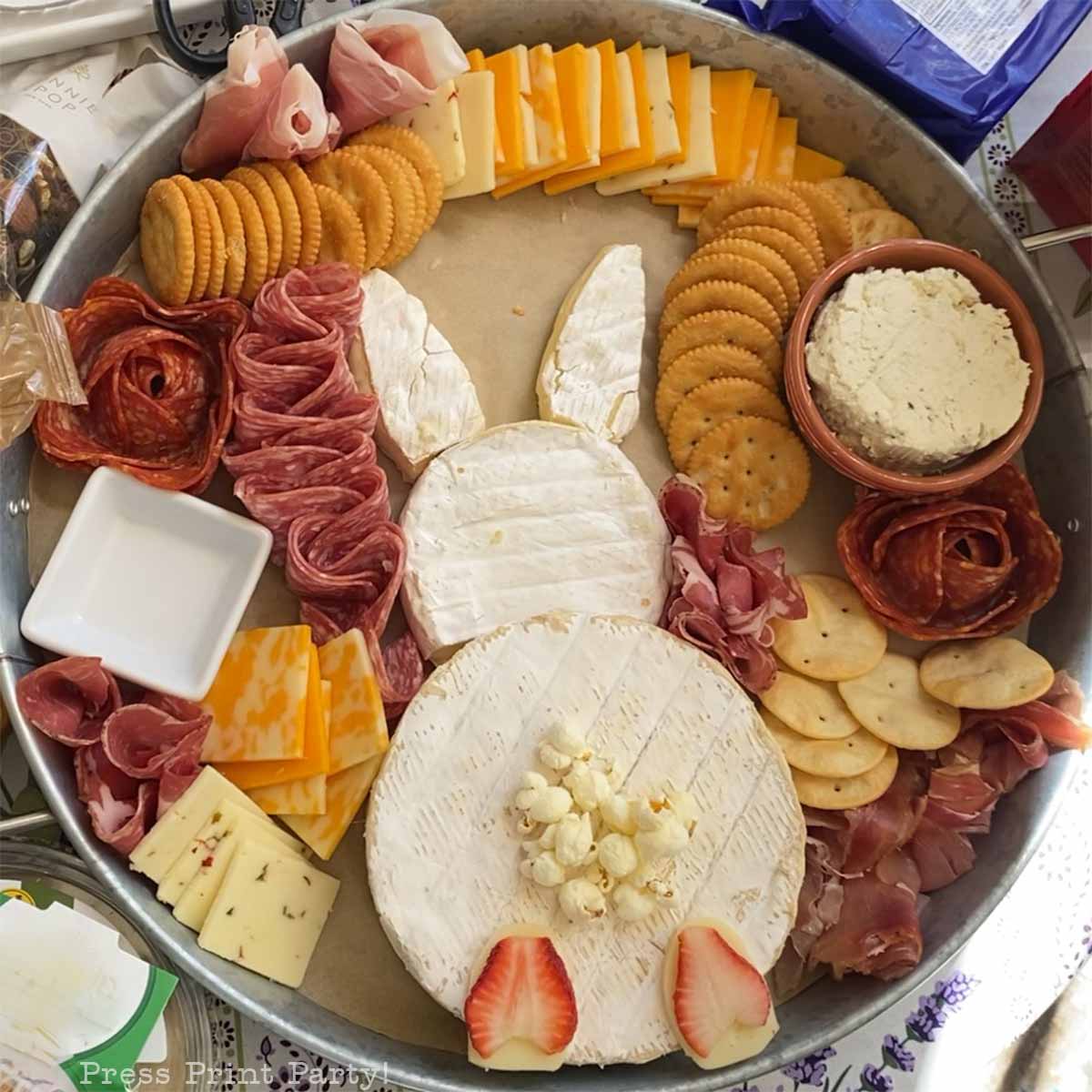 An Easter Bunny Charcuterie Board arranged with round cheeses as the head and body, cheese slices for ears, strawberry feet, popcorn tail, and surrounded by crackers, meats, and extra cheeses for a festive spring treat.