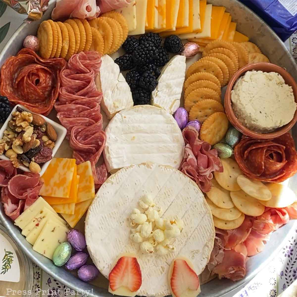 An Easter Bunny Charcuterie Board arranged with circular cheeses for the face and body, cheese ears, popcorn as a tail, plus berries, crackers, meats, nuts, chocolate eggs, and assorted cheeses all around.