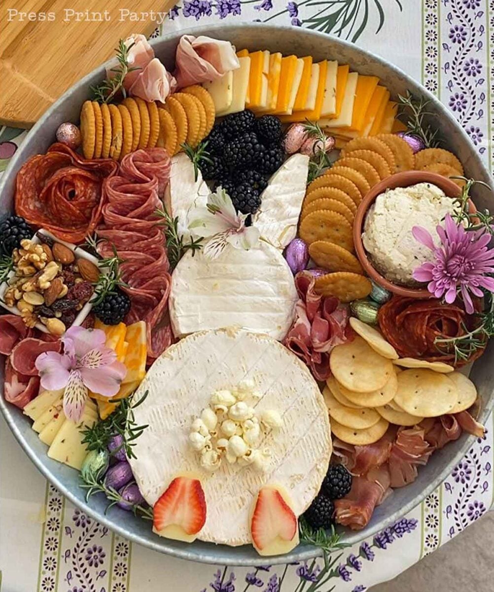An Easter Bunny Charcuterie Board arranged in a round tray with cheeses, crackers, salami, berries, nuts, and dip. Items are creatively shaped to form a bunny and decorated with flowers and herbs on a patterned tablecloth.