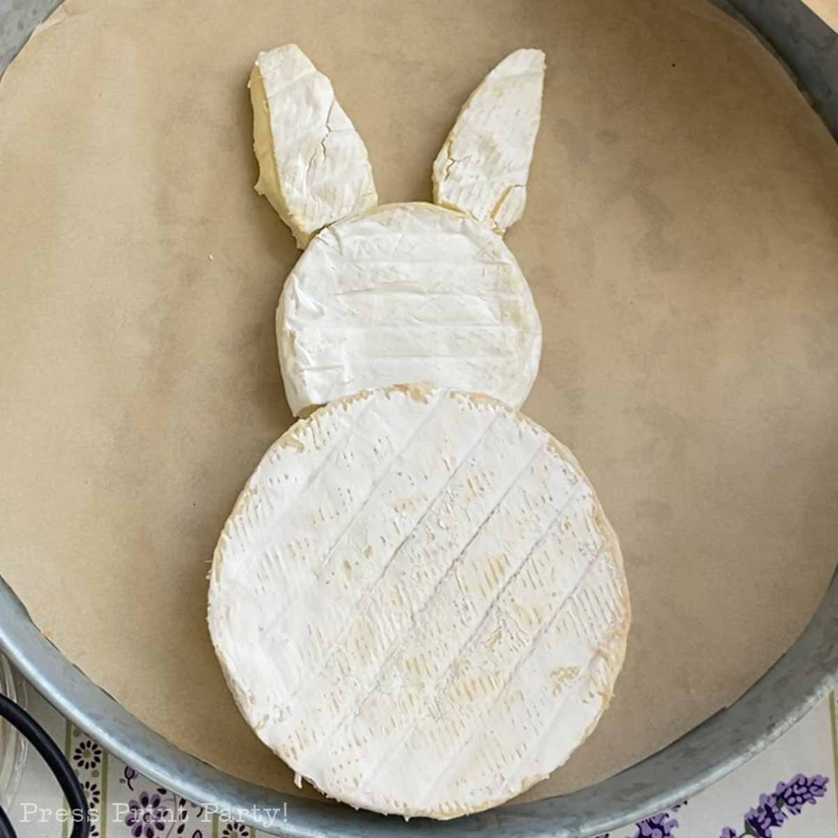 A round wheel of brie cheese with a smaller oval piece and two ear-shaped pieces on top, arranged to resemble a bunnyβperfect as the centerpiece for your Easter Bunny Charcuterie Board on parchment paper.