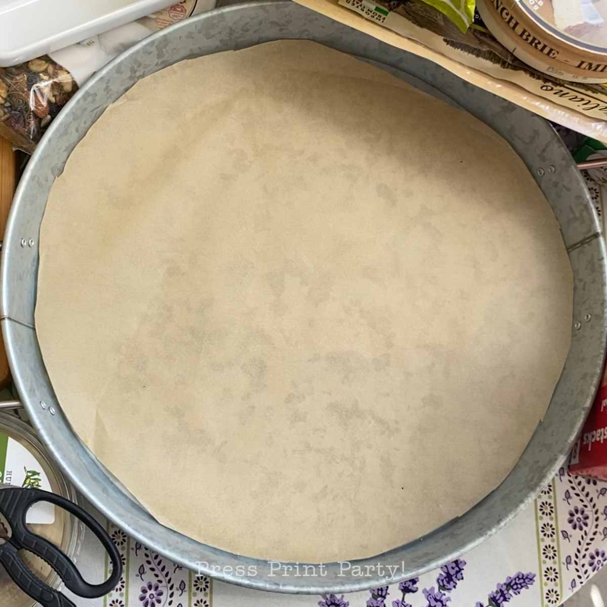 A round baking pan lined with parchment paper sits on a cluttered surface, surrounded by scissors, packaging, and hints of preparation for an Easter Bunny Charcuterie Board.