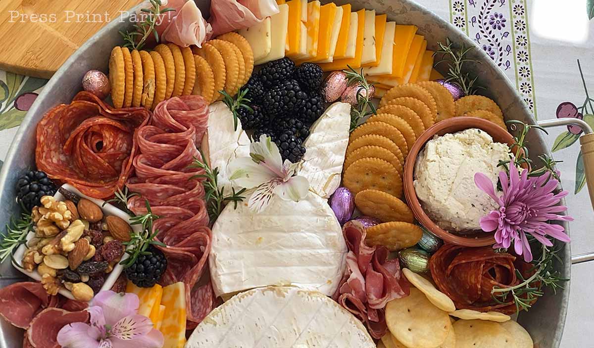 A round Easter Bunny Charcuterie Board features crackers, sliced cheese, meats, blackberries, nuts, and a soft cheese spread. At the center is Brie cheese shaped like a bunny, garnished with flowers and herbs for festive decoration.